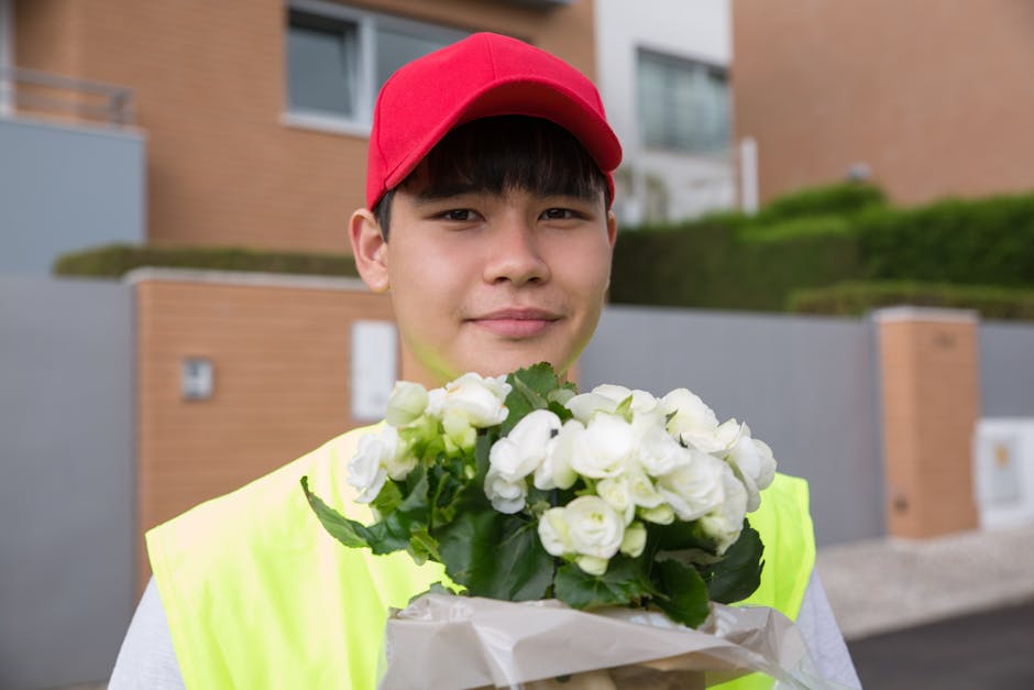 A young man with fair skin, wearing a red cap and a high-visibility yellow vest, is standing outdoors in front of modern residential buildings with brown brick and grey concrete facades. He is holding