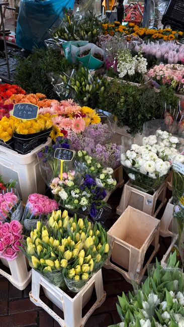 A vibrant display of freshly cut flowers at a local flower market, featuring bouquets of yellow tulips in white plastic crates, pink roses, white ranunculus, purple and white daisies, and assorted gre