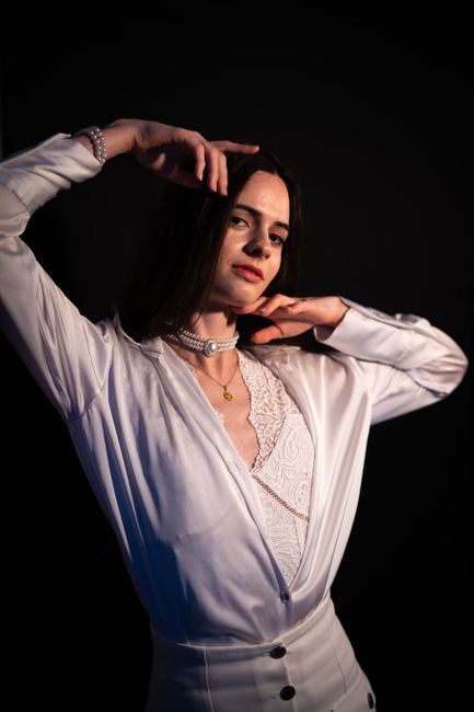 A woman with shoulder-length dark hair, dressed in a white satin blouse with lace details, stands against a dark background. She is posing with her right hand raised to her head and her left hand rest