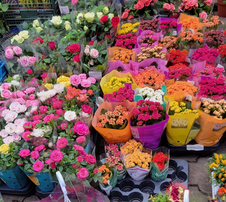 A vibrant display of fresh flowers including pink, white, and yellow roses, as well as various small, colorful blooms arranged in bouquets wrapped in pink, purple, orange, and yellow paper. The flower