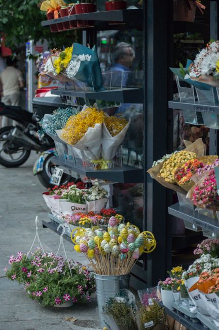 An outdoor flower display at a local florist featuring a variety of vibrant floral arrangements and bouquets arranged on metal shelves. The top shelf holds a bouquet of yellow daffodils and purple hya