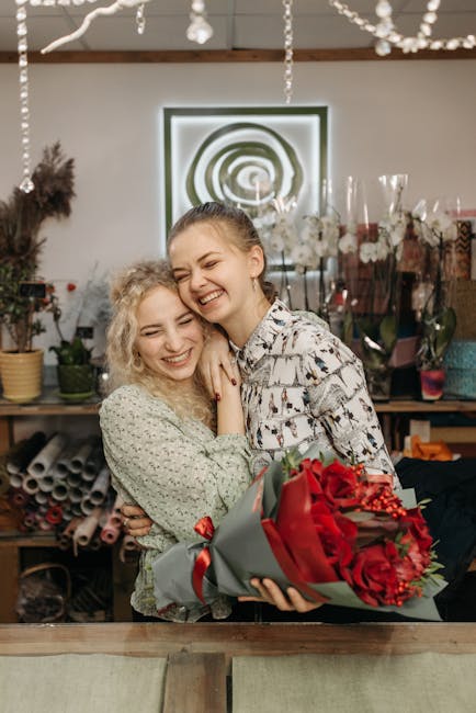 Two young women are sharing a joyful moment in a flower shop, with one holding a large, vibrant bouquet of red roses wrapped in gray and red paper, accented with red berries. The bouquet features fres