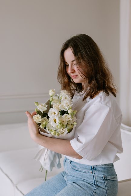 A young woman with shoulder-length brown hair, wearing a white blouse and light blue jeans, is sitting indoors on a cream-colored sofa. She is gently holding a bouquet of fresh, predominantly white fl